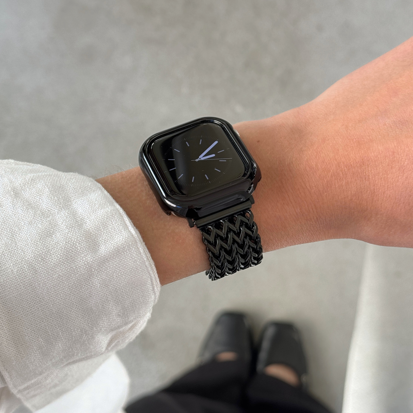 A close-up of a person's wrist wearing a black Apple smartwatch with a black mesh band and a matching bezel, set against a gray concrete background with feet visible at the bottom.