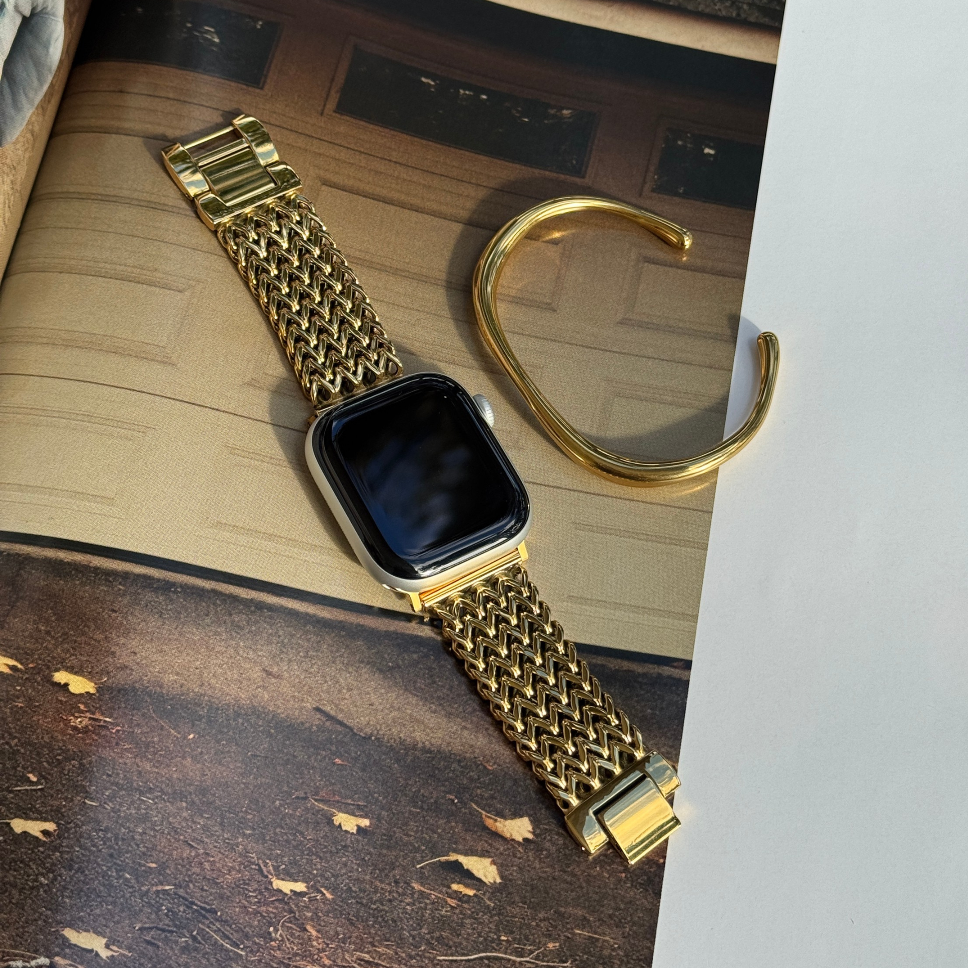A gold-tone metal mesh watch band attached to an Apple smartwatch, displayed diagonally on a textured surface next to a matching gold cuff bracelet.
