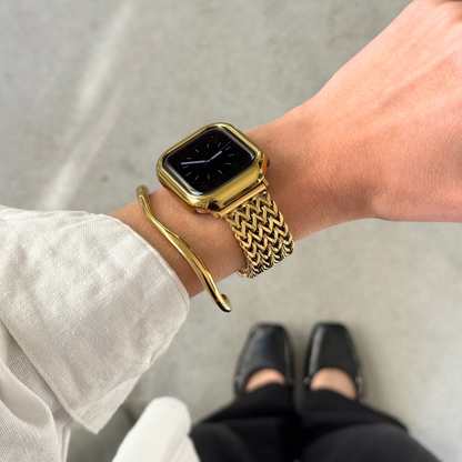 A close-up of a person's wrist wearing a gold-toned Apple smartwatch with a woven mesh band, along with a slim gold bangle bracelet and a gold matching bezel. The photo is taken from above, showing the person's arm, hand, white sleeve, black pants, and black shoes against a gray floor.