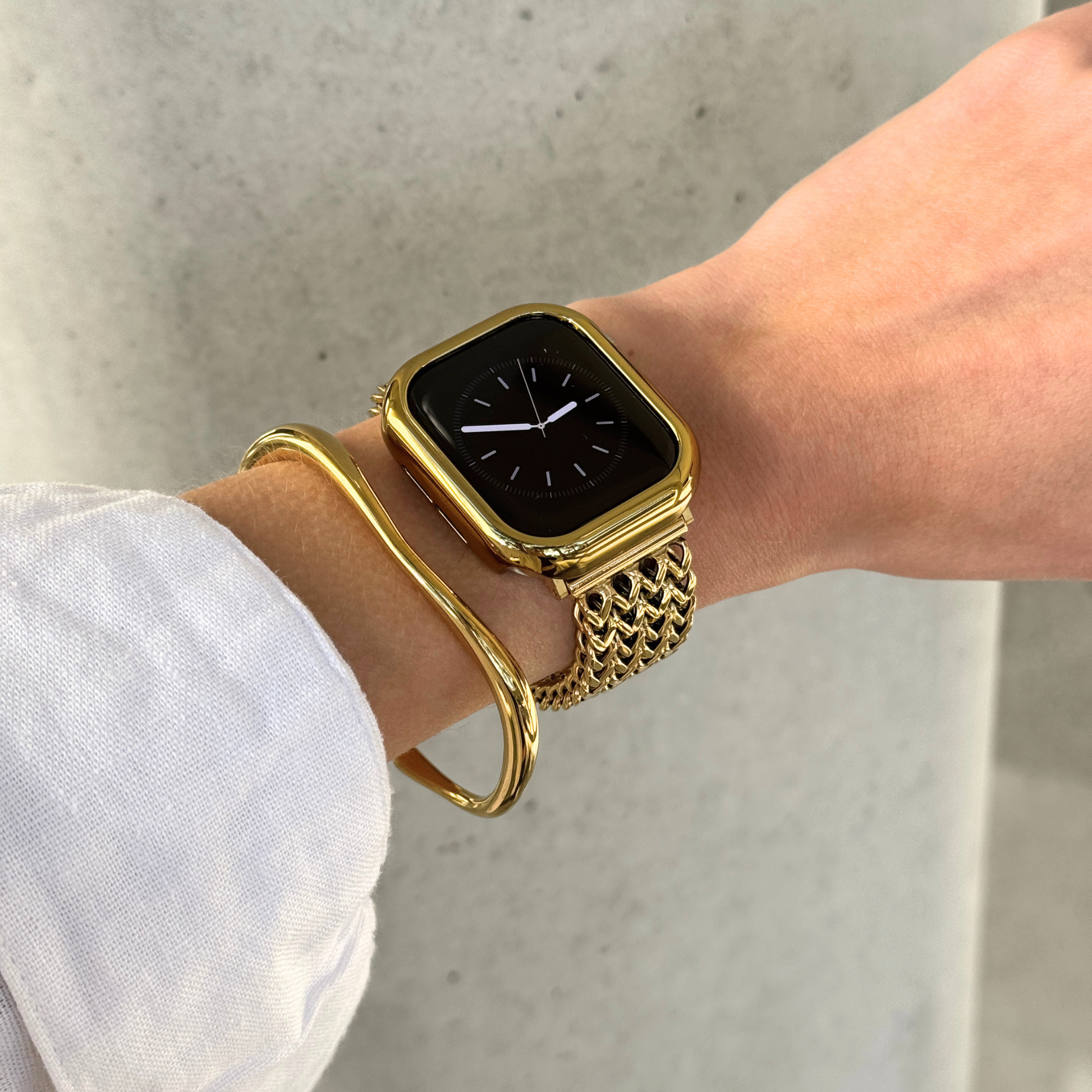 A close-up of a person's wrist wearing a gold-toned Apple watch with a textured gold link band, along with a matching gold bangle bracelet and a matching watch case cover.