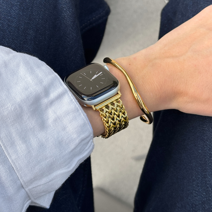 A close-up of a person's wrist wearing a gold mesh metal watch band on an Apple smartwatch, along with a gold bangle bracelet, a white shirt cuff, and dark blue jeans.