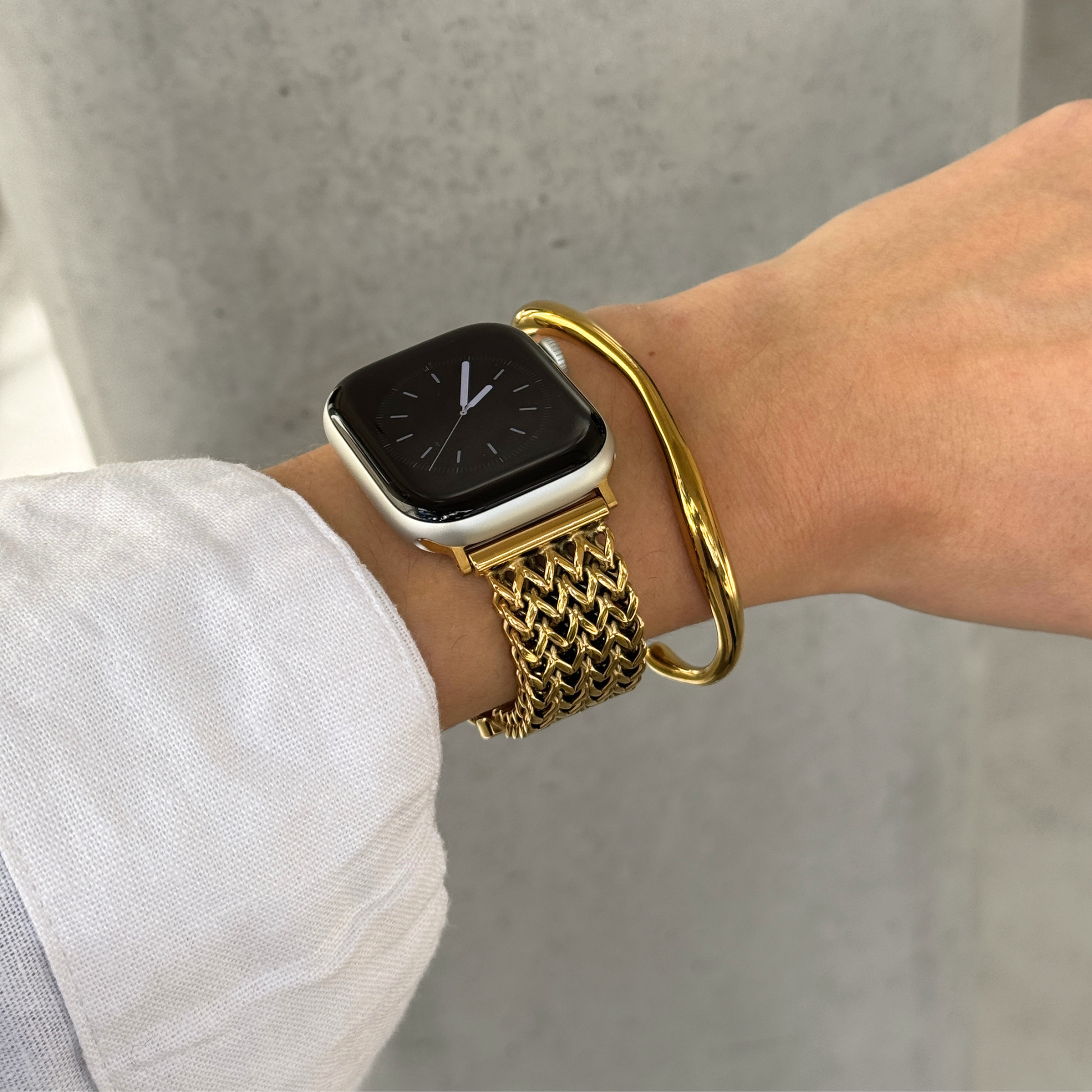 A close-up of a person's wrist wearing a gold mesh watch band on an Apple Watch, paired with a gold bangle bracelet, against a gray background.