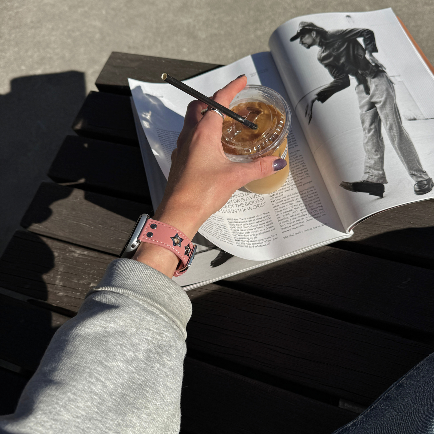A person's hand holding an iced coffee over an open magazine on a dark wooden slatted table. The hand is wearing a pink Apple Watch faux-leather band with silver star details.