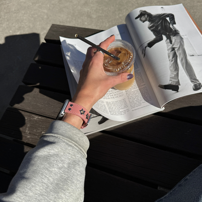 A person's hand holding an iced coffee over an open magazine on a dark wooden slatted table. The hand is wearing a pink Apple Watch faux-leather band with silver star details.