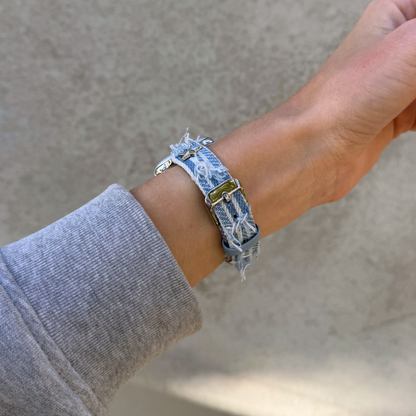 A close-up of a person's wrist wearing a light blue denim Apple Watch bracelet with distressed frayed edges and a silver-toned buckle, paired with a gray long-sleeve shirt against a neutral concrete background.