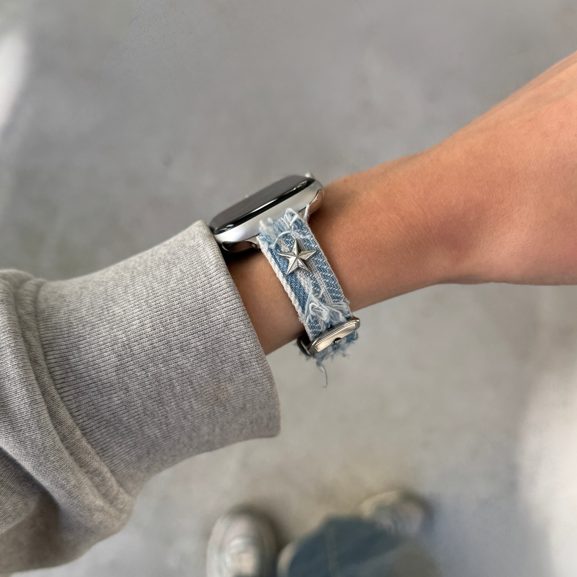 A close-up of a person's wrist wearing a light blue denim Apple Watch strap with a star charm, paired with a gray sweatshirt sleeve, against a gray concrete floor background.