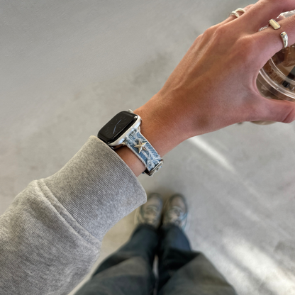 A person wearing a light blue denim Apple watch strap with a star pattern on a smartwatch, shown from a first-person perspective while holding an iced coffee. The person is wearing a gray sweatshirt, dark jeans, and sneakers, with silver rings on their fingers.