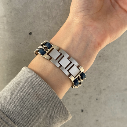 A close-up of a person's wrist wearing a silver-toned metal link Apple Watch bracelet with dark blue denim braided cord accents, paired with a gray long-sleeve shirt against a concrete background.