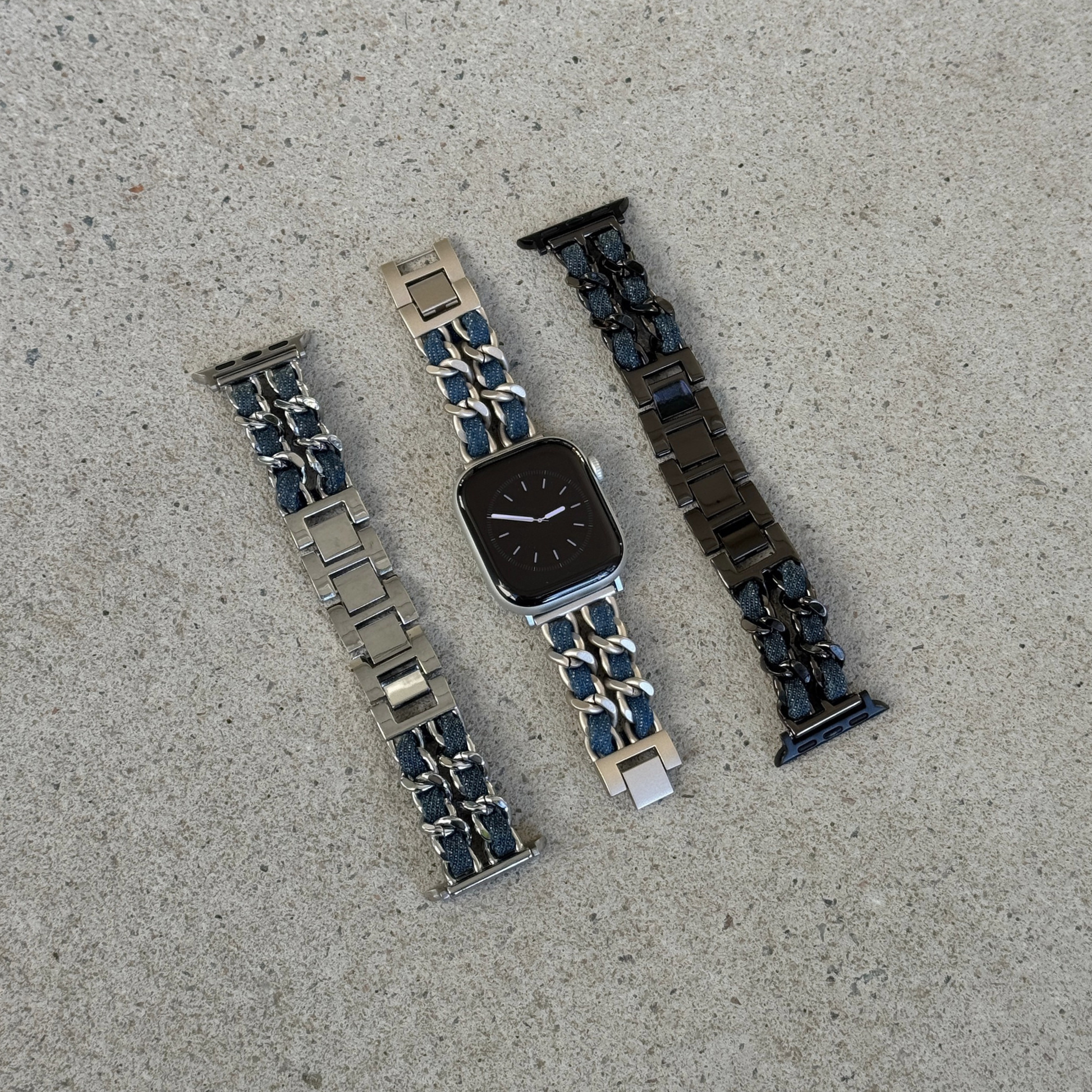 Three metal watch bands arranged on a light gray concrete surface, on a silver Apple watch. The left band is silver with dark blue denim accents, the center band is starlight with dark blue denim accents, and the right band is black with dark blue denim accents.
