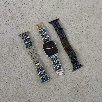 Three metal watch bands arranged on a light gray concrete surface, on a silver Apple watch. The left band is silver with dark blue denim accents, the center band is starlight with dark blue denim accents, and the right band is black with dark blue denim accents.
