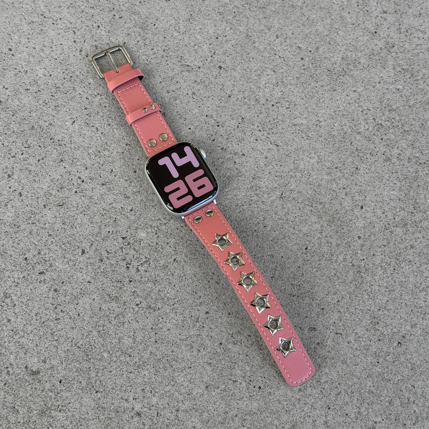 A pink leather watch band with silver star-shaped studs, shown attached to an Apple Watch on a textured gray concrete surface.