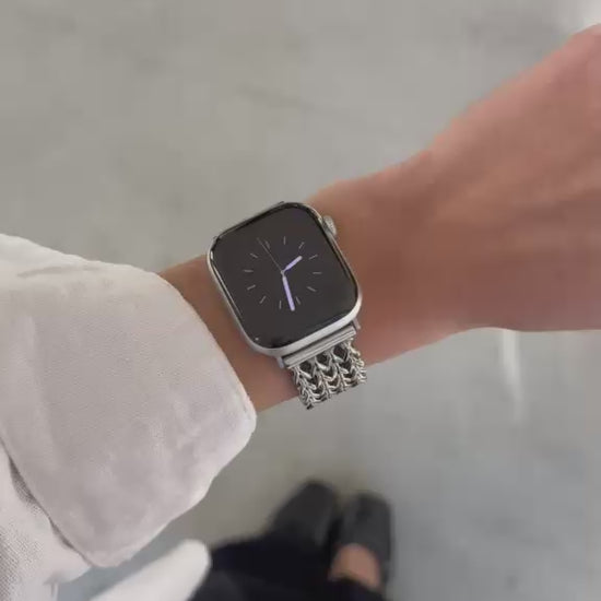 A close-up video of a person's hand wearing a silver mesh Apple Watch band with a silver Apple Watch, set against a modern concrete pillar background. 