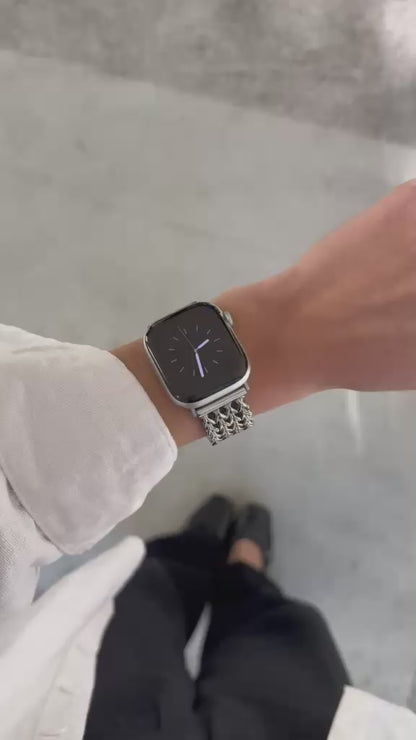 A close-up video of a person's hand wearing a silver mesh Apple Watch band with a silver Apple Watch, set against a modern concrete pillar background. 