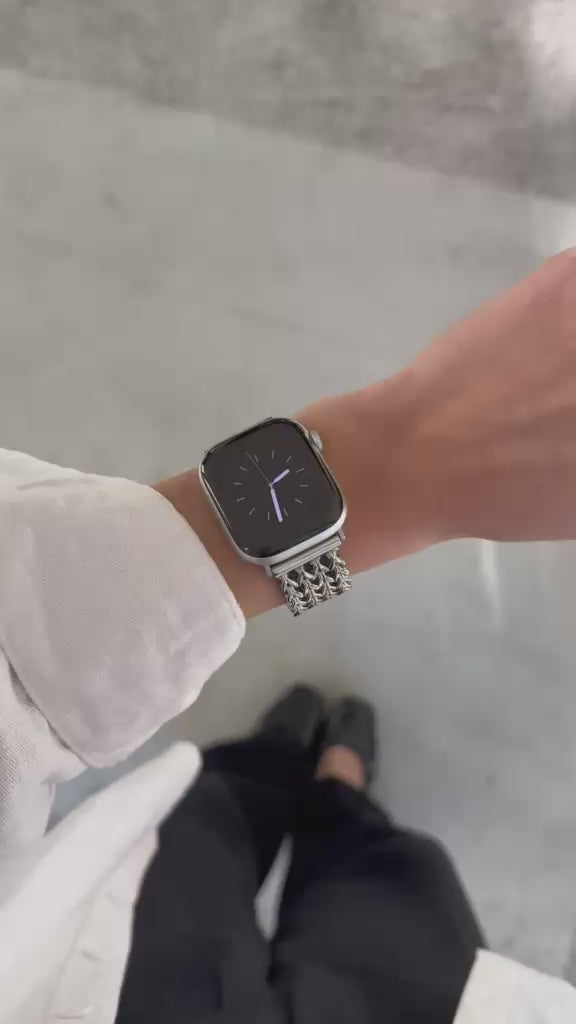 A close-up video of a person's hand wearing a silver mesh Apple Watch band with a silver Apple Watch, set against a modern concrete pillar background. 