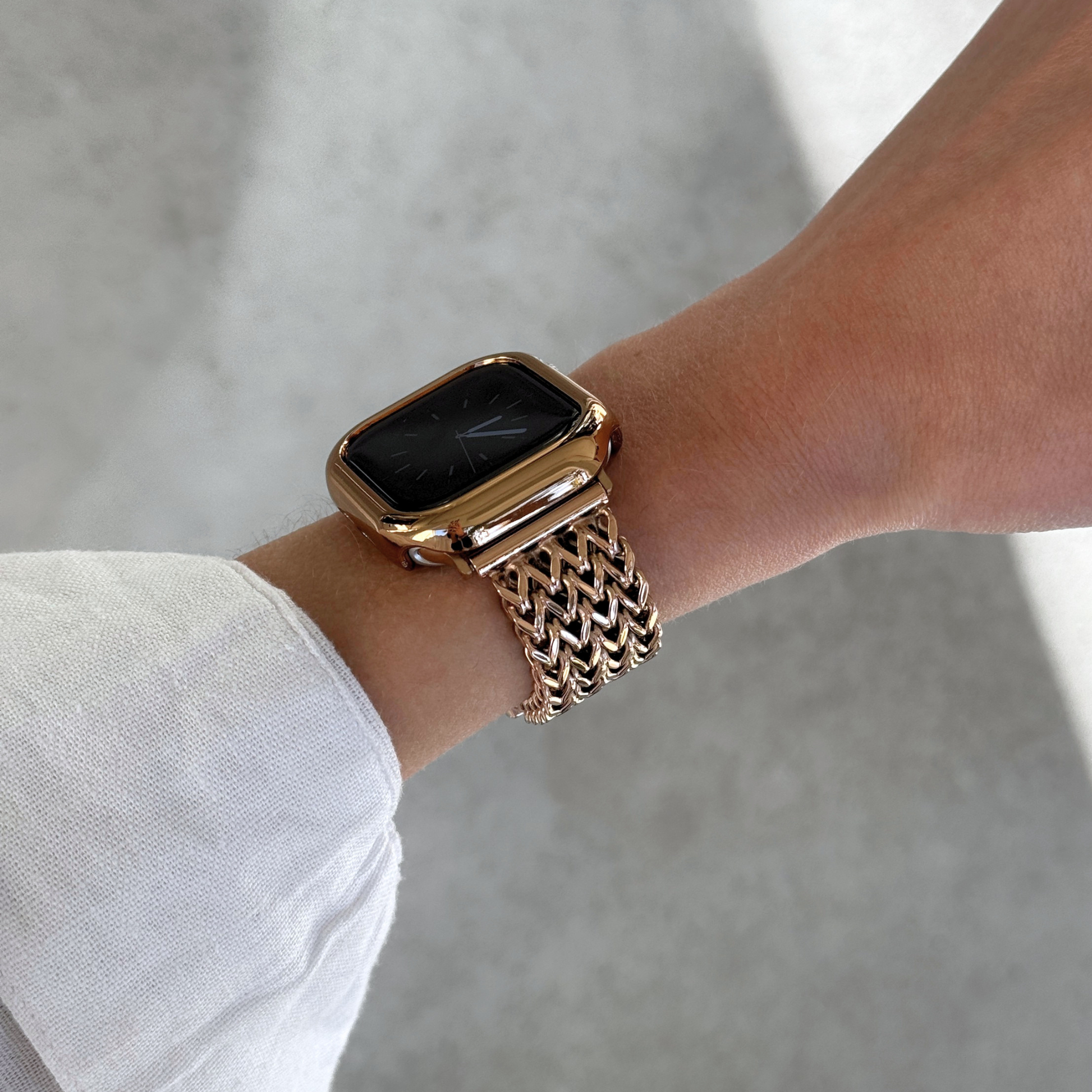 A close-up of a person's wrist wearing a rose gold-toned Apple smartwatch with a matching metallic mesh band and bezel, set against a soft gray concrete background.