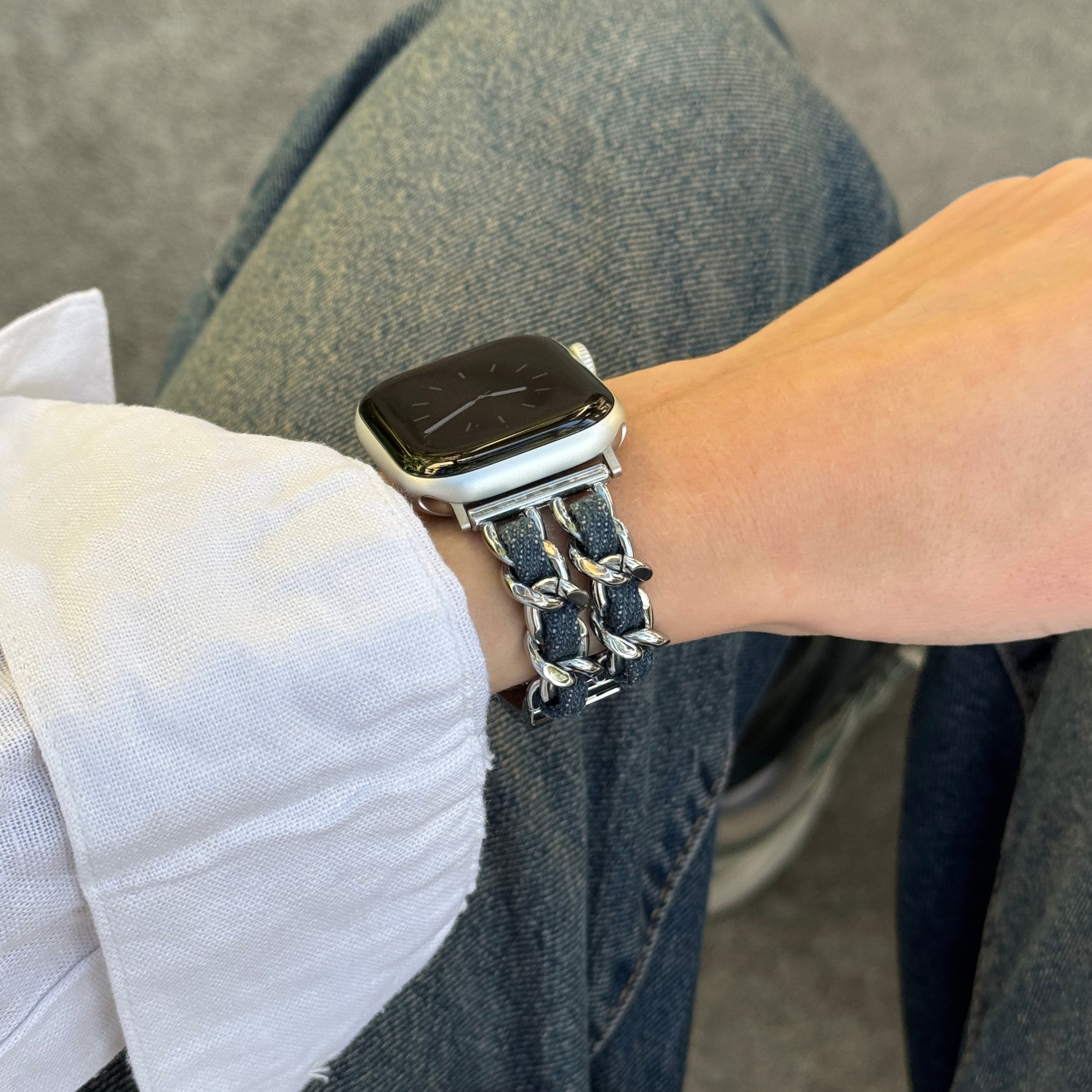 A close-up of a person's wrist wearing a silver-toned metal Apple watch band with dark woven accents, paired with a smartwatch, blue jeans, and a white shirt.