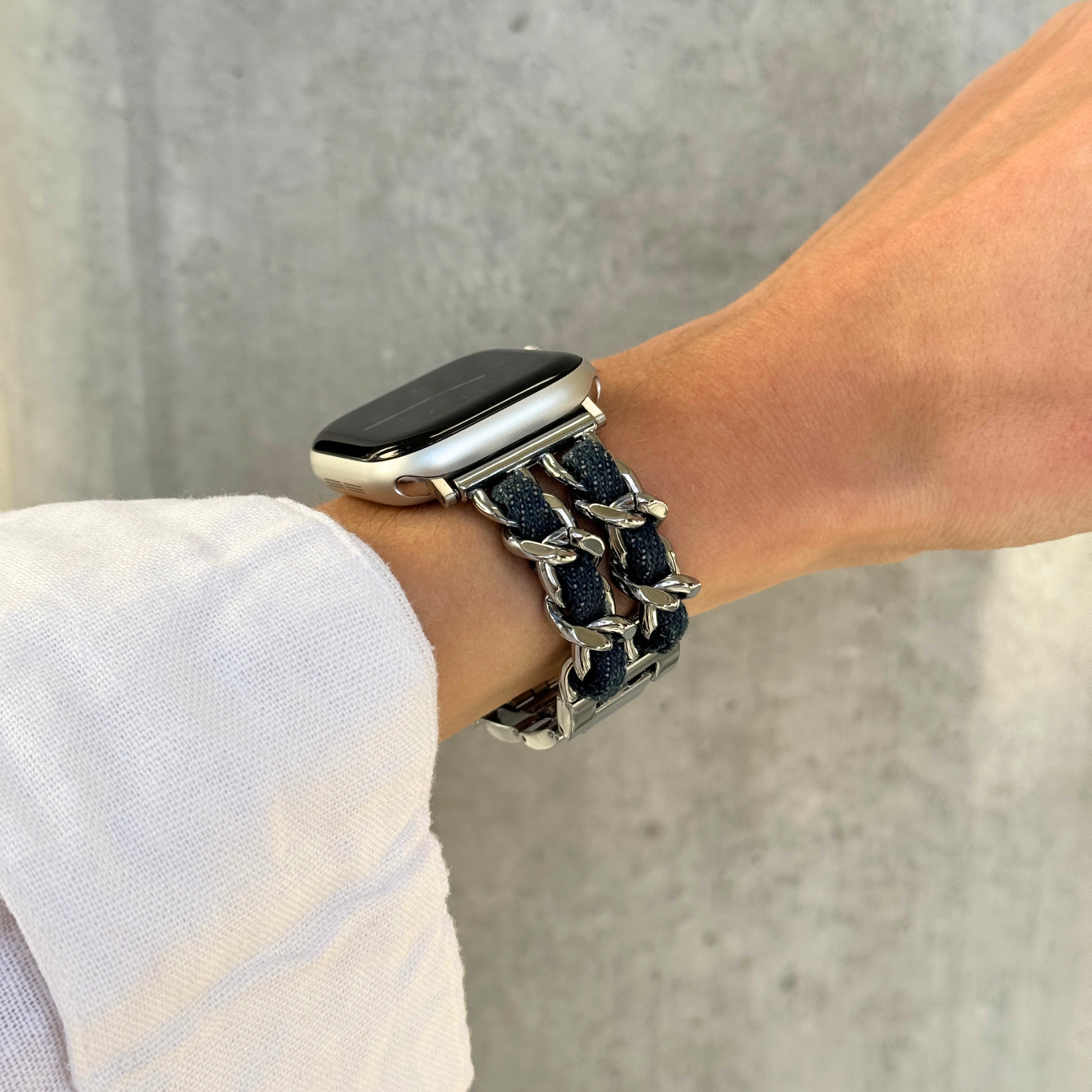 A close-up of a person's wrist wearing a silver-toned metal link bracelet watch band with dark blue woven fabric details, attached to an Apple Watch.