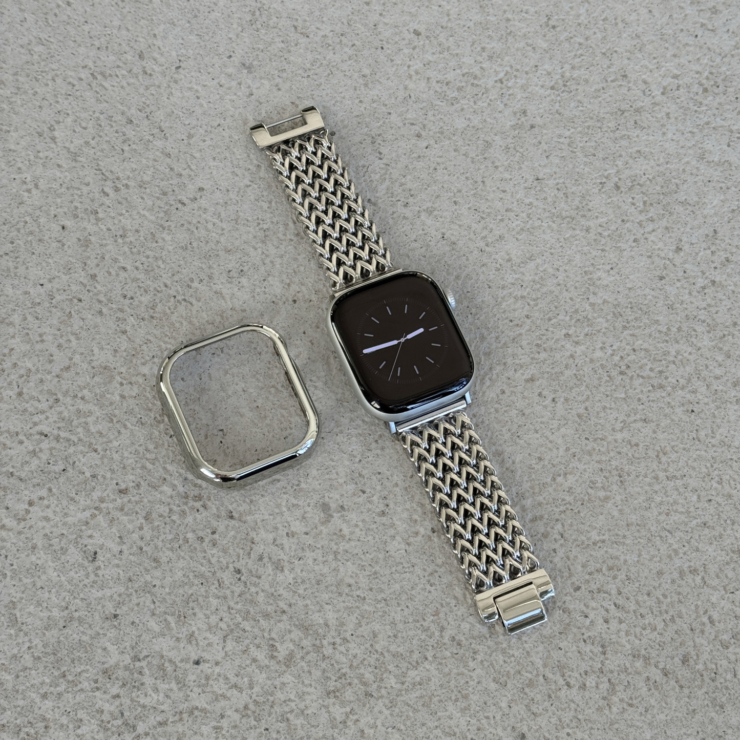 A silver-toned metal mesh watch band with a matching bezel protector, shown laid flat on a textured gray surface next to a smartwatch.