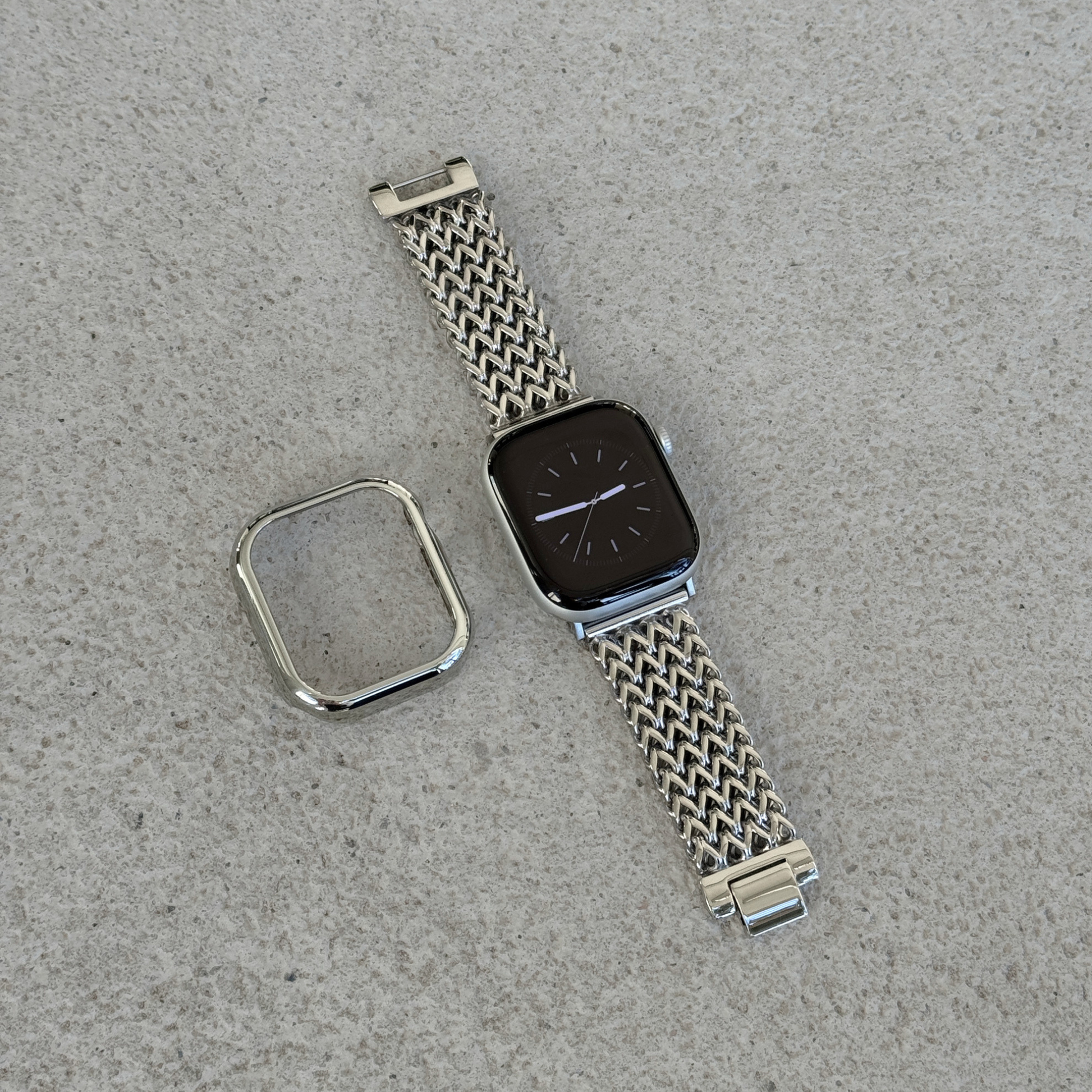 A silver-toned metal mesh watch band with a matching bezel protector, shown laid flat on a textured gray surface next to a smartwatch.