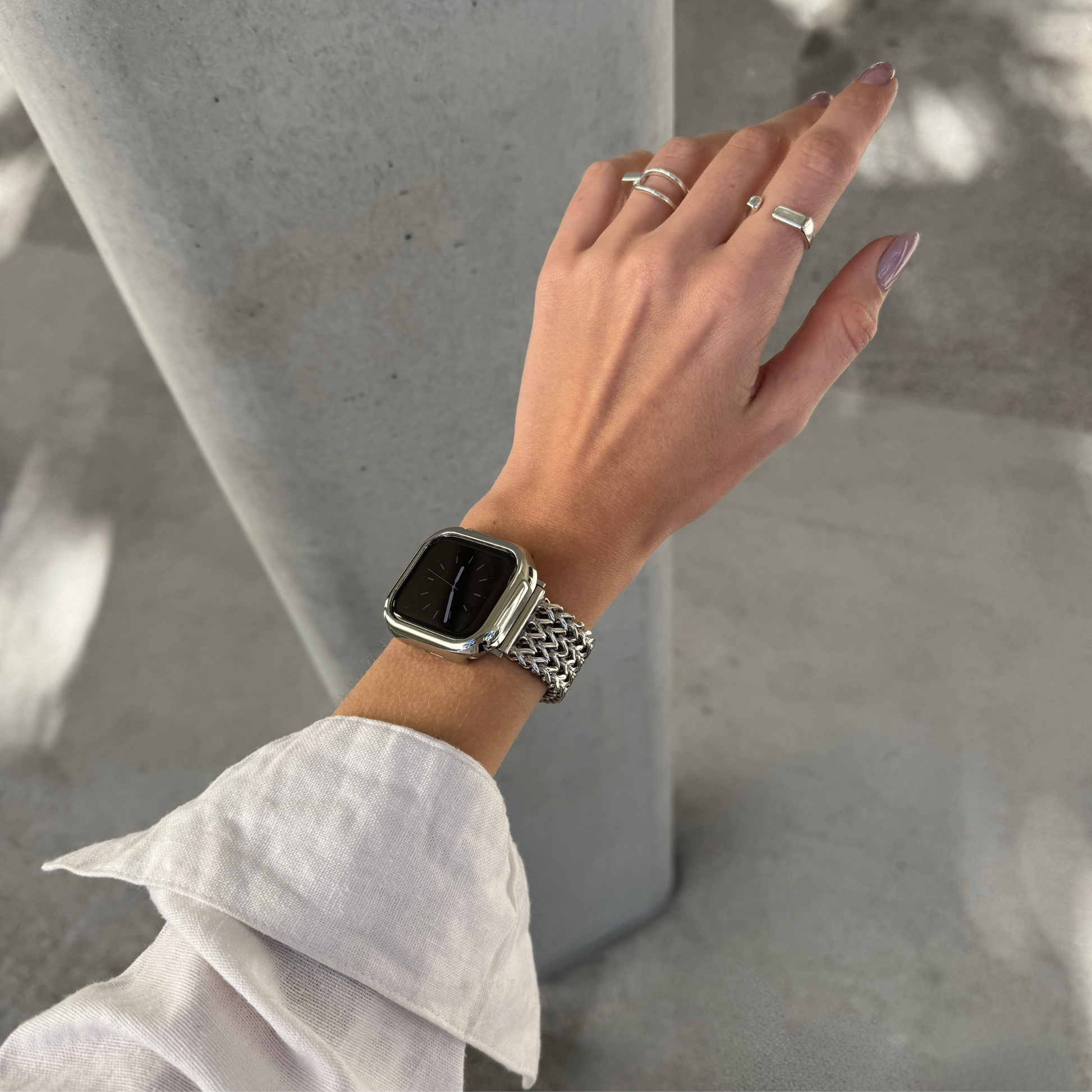 A close-up of a person's hand and wrist wearing a silver Apple Watch with a silver mesh band and silver watch case cover, along with silver rings, set against a concrete pillar background.