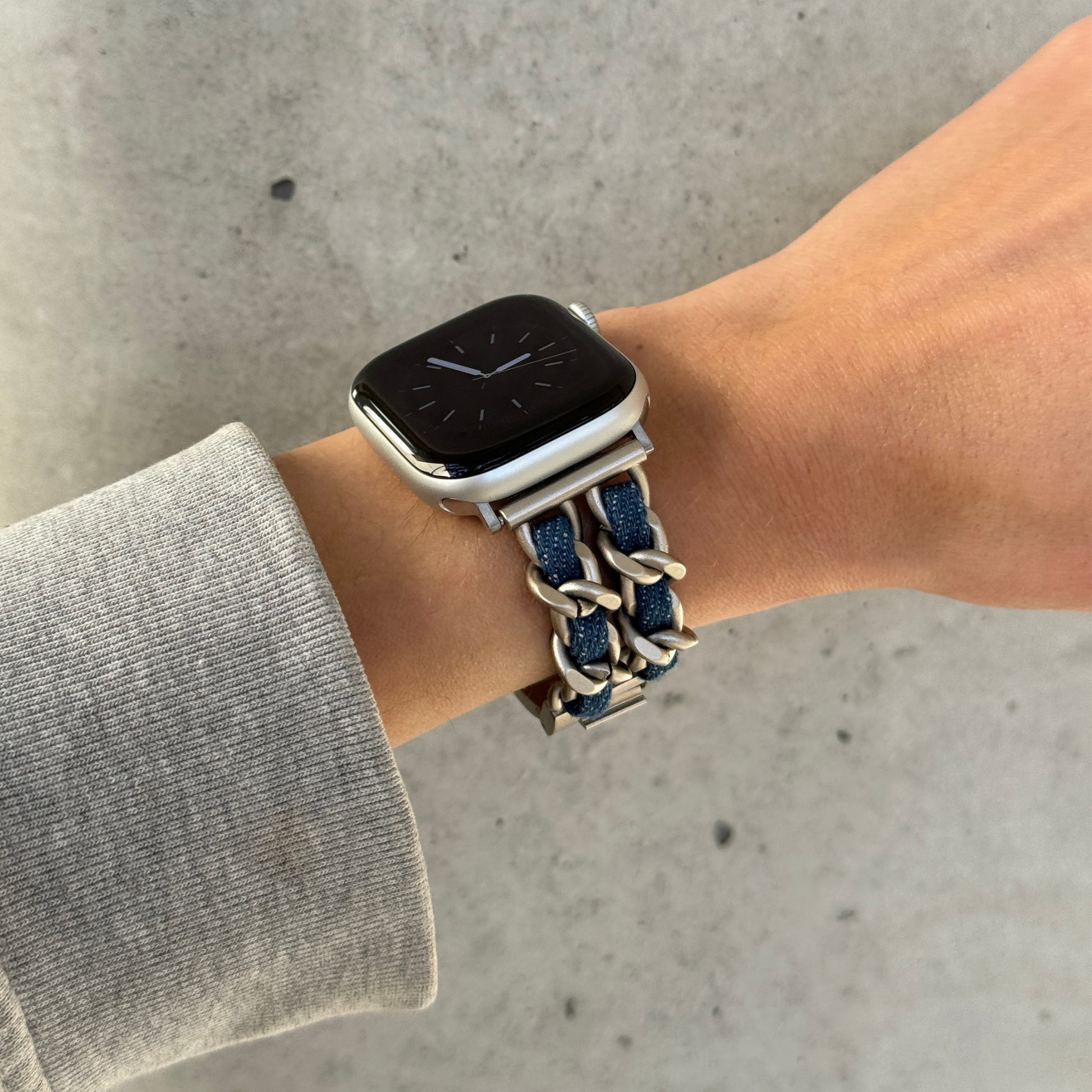 A close-up of a person's wrist wearing a silver Apple smartwatch with a braided denim blue and silver chain strap, set against a gray concrete background.