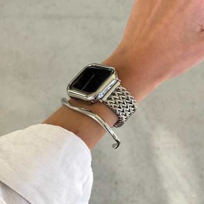 A close-up of a person's wrist wearing a silver Apple watch with a woven metal mesh band, a silver curved bangle bracelet and a matching watch case cover