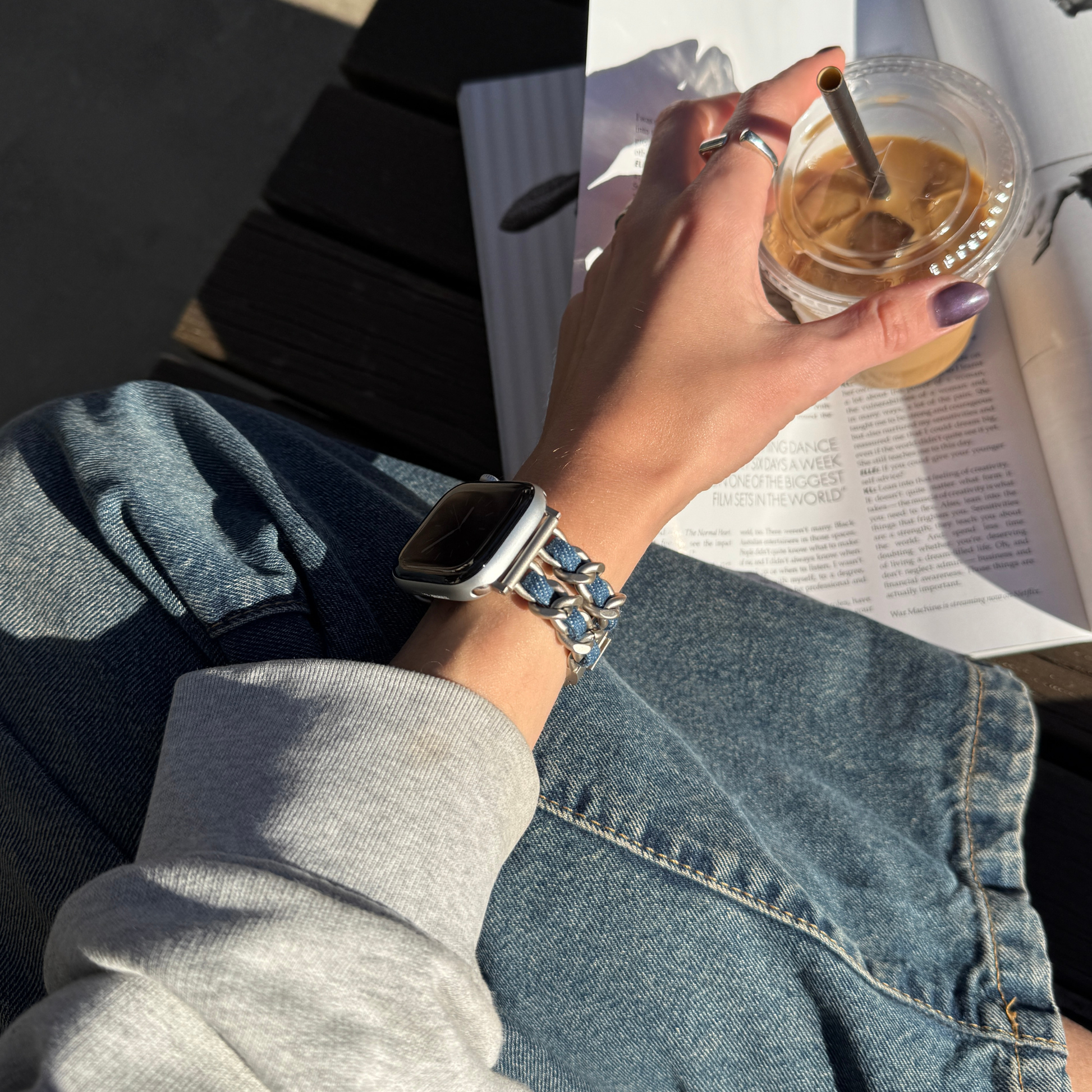 A close-up lifestyle shot of a person wearing a silver chain-link Apple smartwatch band on their wrist, sitting outdoors next to an open book and holding an iced coffee.