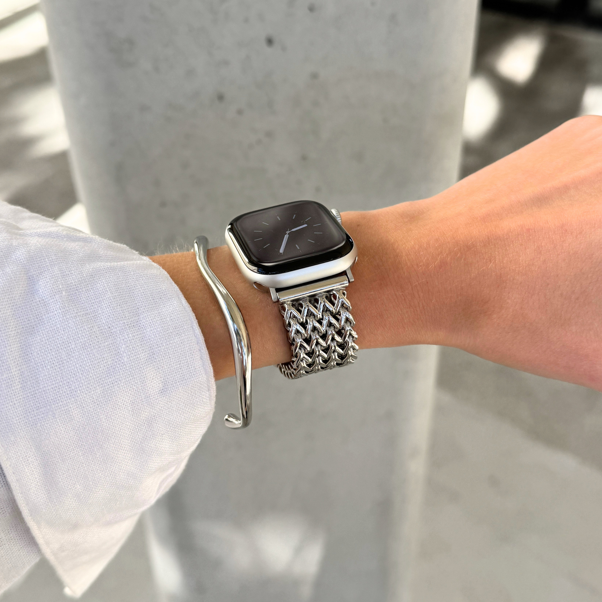 A close-up of a person's wrist wearing a silver mesh Apple watch band, with a silver bangle bracelet also visible, set against a blurred concrete background.