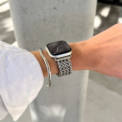 A close-up of a person's wrist wearing a silver mesh Apple watch band, with a silver bangle bracelet also visible, set against a blurred concrete background.