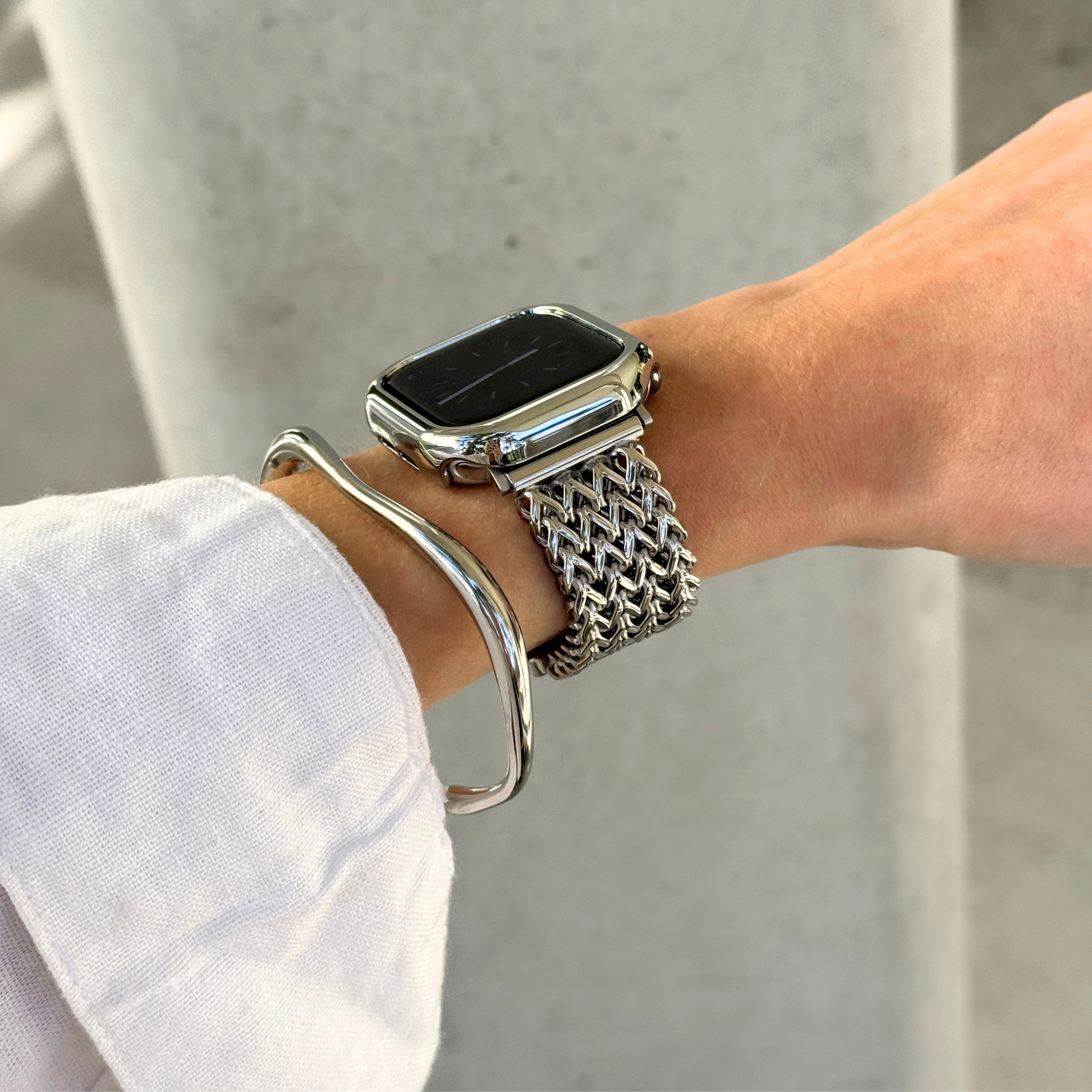 A close-up of a person's wrist wearing a silver mesh Apple watch band with a smartwatch, along with a silver bangle bracelet and a matching bezel, against a light gray concrete background.