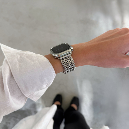 A close-up of a person's wrist wearing a silver metallic mesh Apple Watch band and a matching bezel cover, with a white shirt sleeve visible and a gray floor in the background.