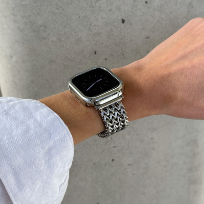 A close-up of a person's wrist wearing a silver smartwatch with a shiny metallic mesh Apple watch band and a matching case cover, set against a gray concrete wall.