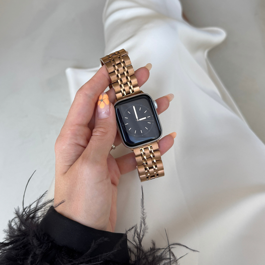 Rose gold link Apple Watch band held in a hand above white satin. Waterproof stainless steel iWatch strap with a secure clasp, compatible with Apple Watch 38mm to 49mm, Series 1-11, SE and Ultra