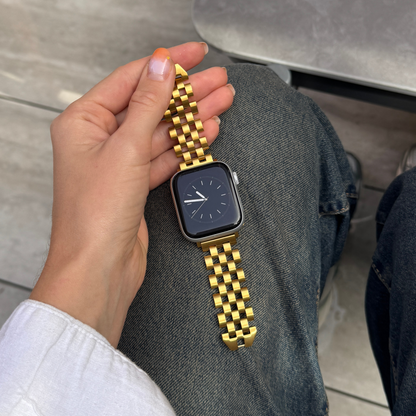 Hand holding a smartwatch with a gold bracelet against a neutral background