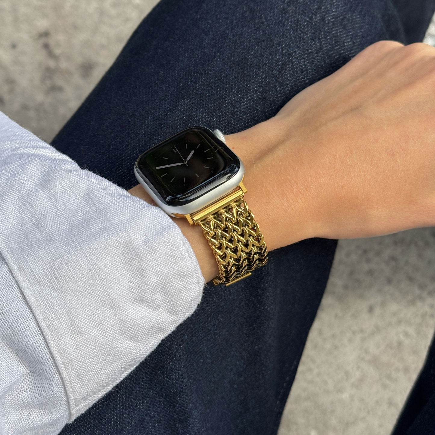 A close-up of a person's wrist wearing a gold-tone mesh metal band on a Apple smartwatch, with dark blue jeans and a light-colored shirt visible in the background
