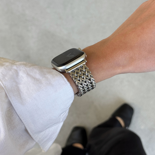A close-up of a person's wrist wearing a silver metal mesh watch band with an Apple Watch. The person is wearing a white long-sleeve shirt and black pants, standing on a light gray concrete floor.