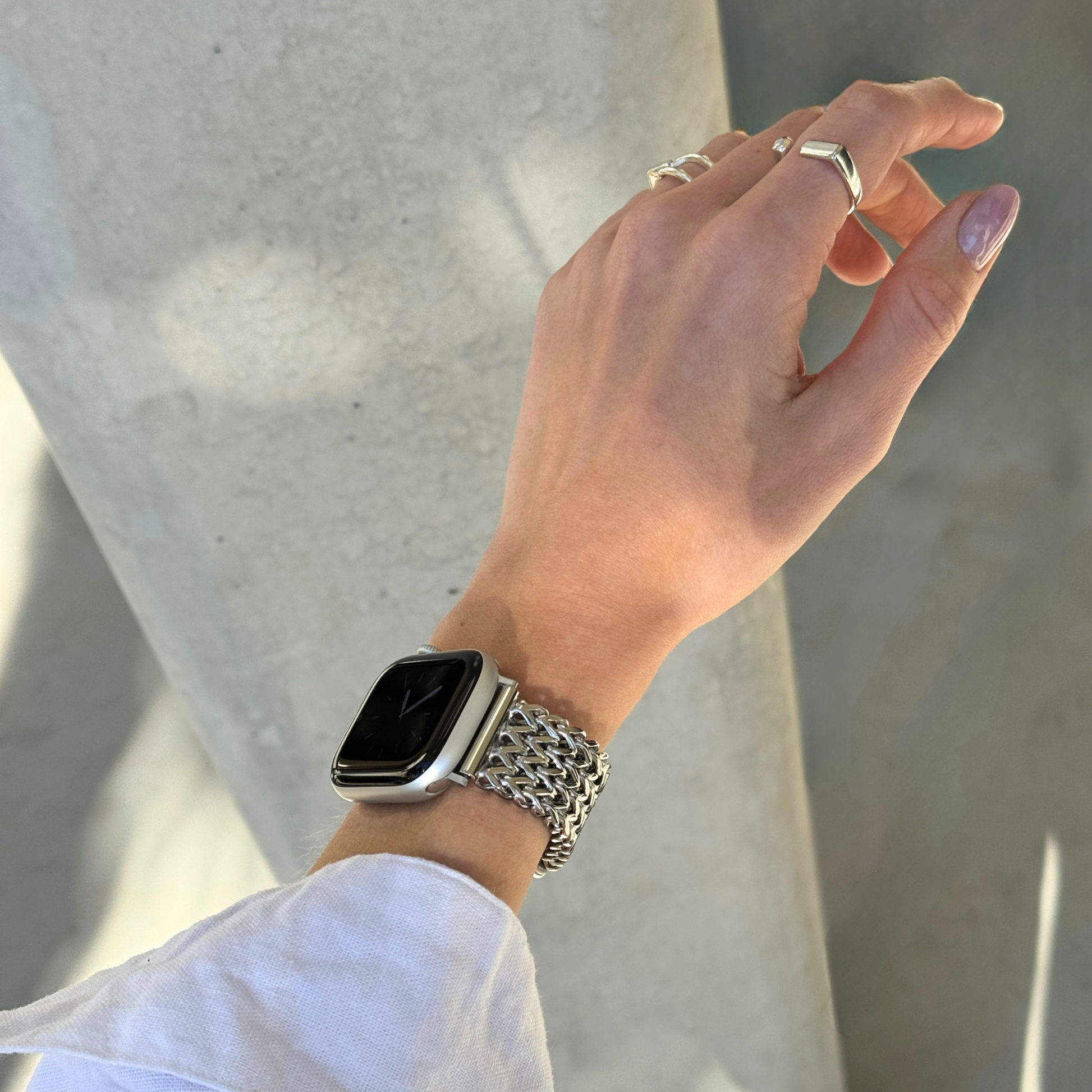 A close-up of a person's hand wearing a silver mesh Apple Watch band with a silver smartwatch, set against a modern concrete pillar background.