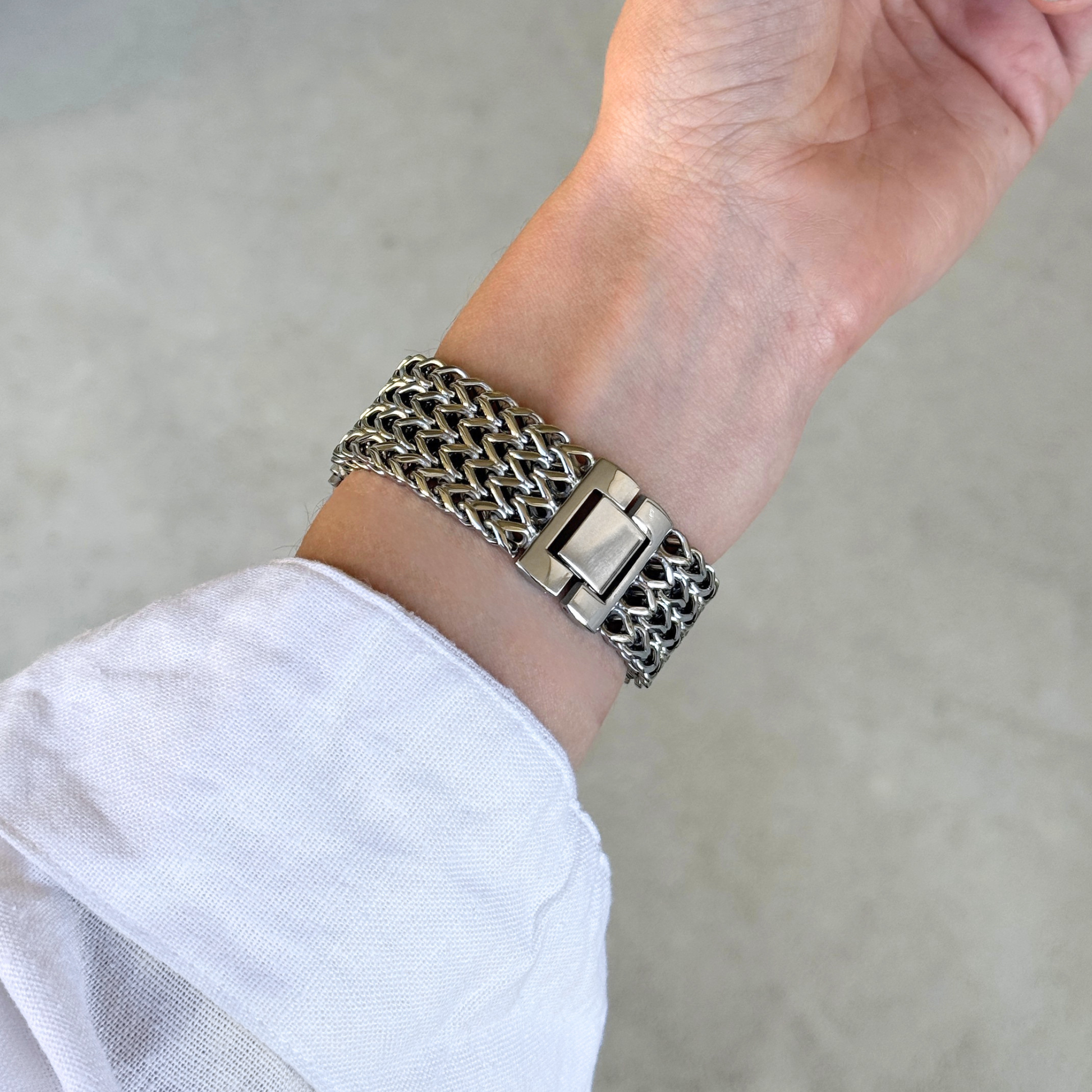 A close-up of a silver-toned metal link bracelet with a wide, textured woven-style Apple Watch band and a rectangular clasp, worn on a person's wrist with a white sleeve visible.
