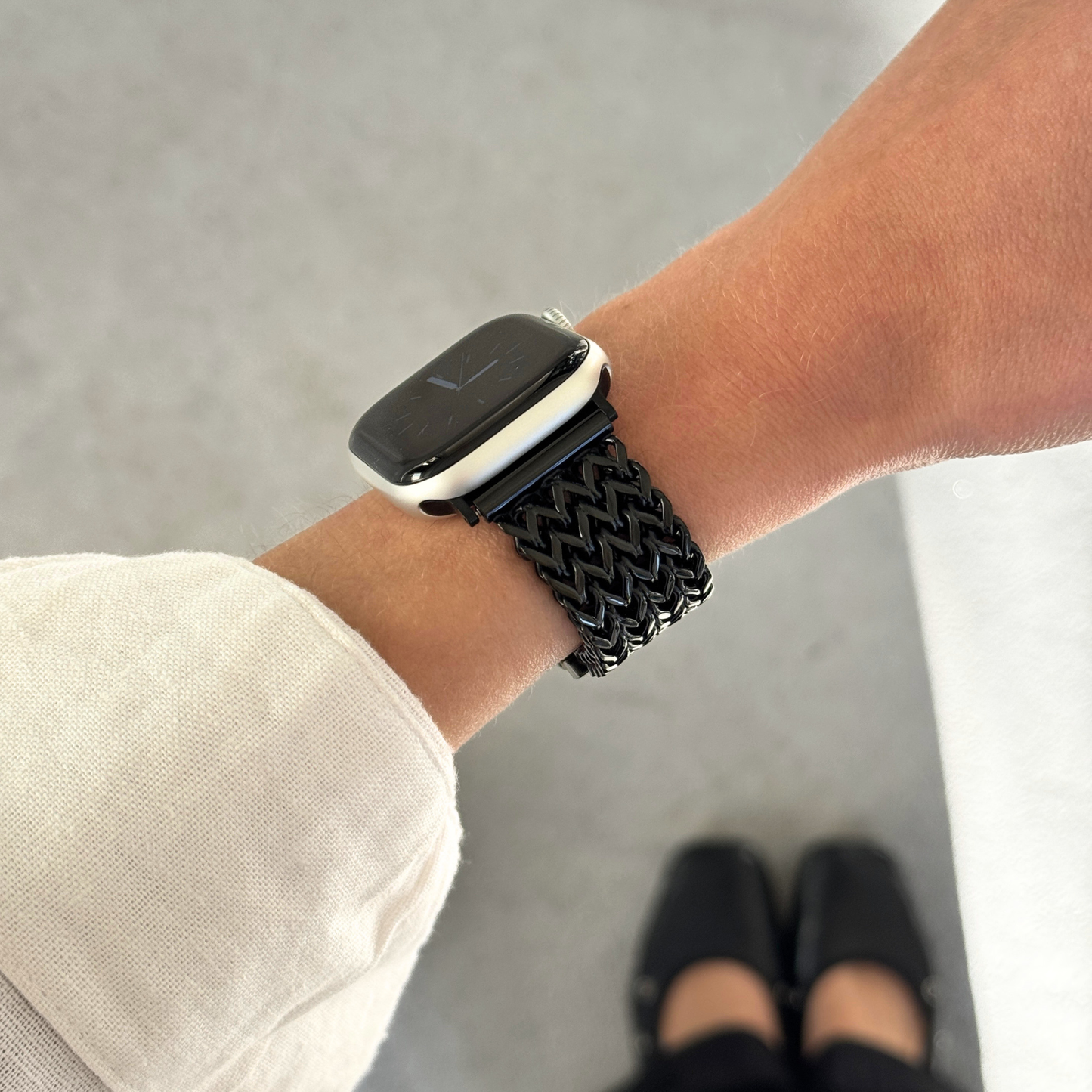 A close-up of a person's wrist wearing a black braided metal mesh band attached to an Apple Watch, with a cream-colored sleeve visible and feet in black flats in the background.