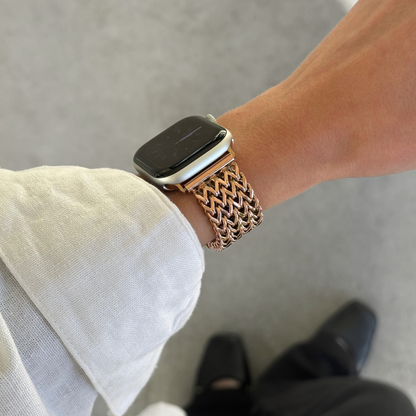 A close-up of a person's wrist wearing a rose gold mesh metal band on an Apple Watch, with a white linen sleeve visible and a gray concrete floor in the background.