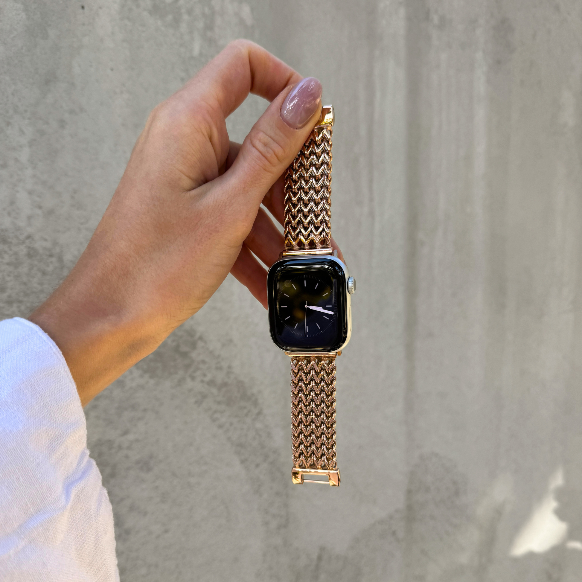 A close-up of a hand holding an Apple Watch with a rose gold mesh metal band against a textured gray concrete wall.