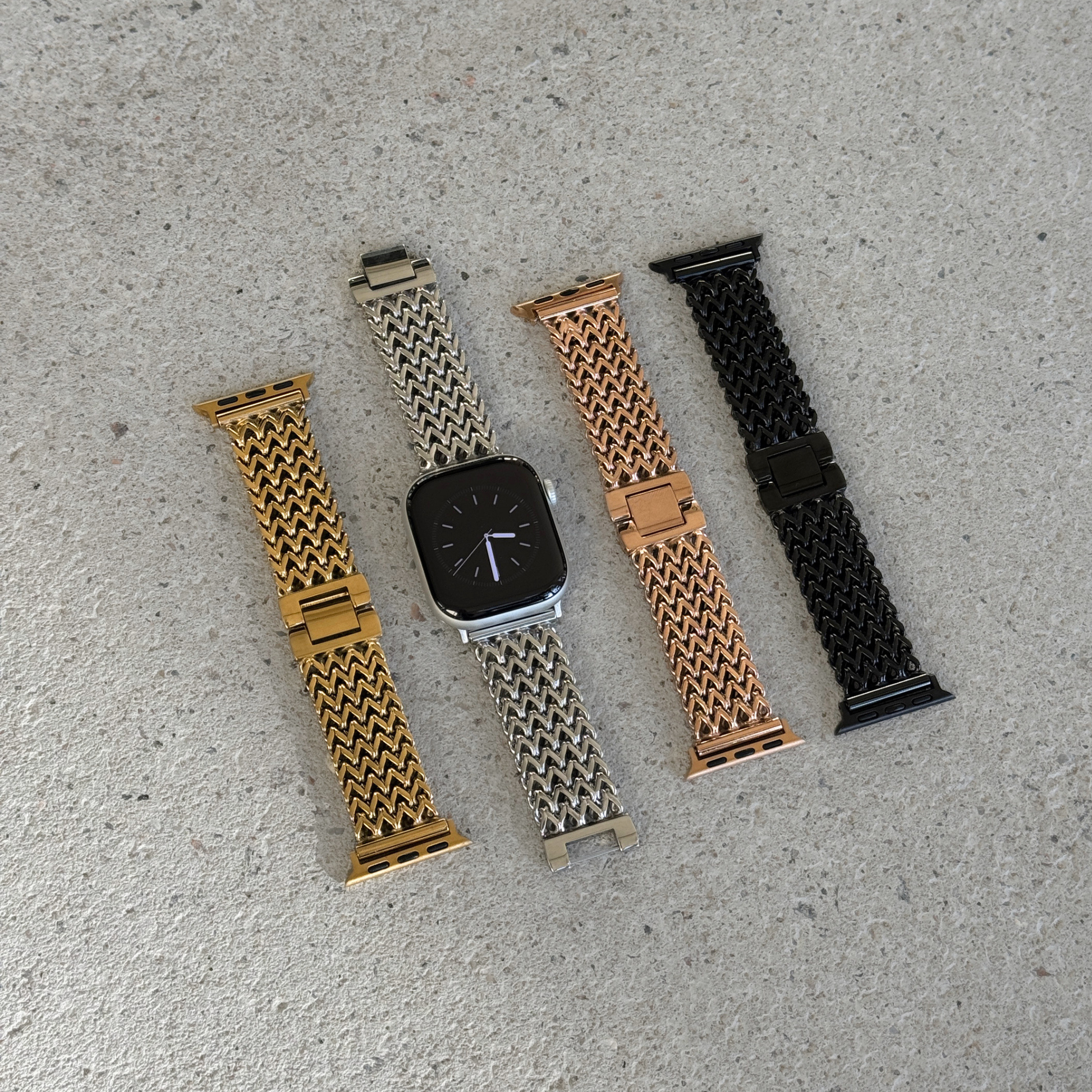Four metal mesh Apple watch straps in gold, silver, rose gold, and black, displayed with silver smartwatches on a textured gray concrete surface.
