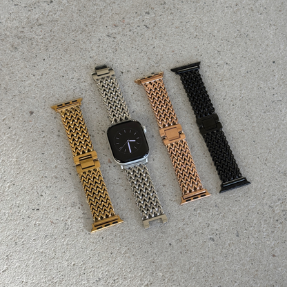Four metal mesh Apple watch straps in gold, silver, rose gold, and black, displayed with silver smartwatches on a textured gray concrete surface.
