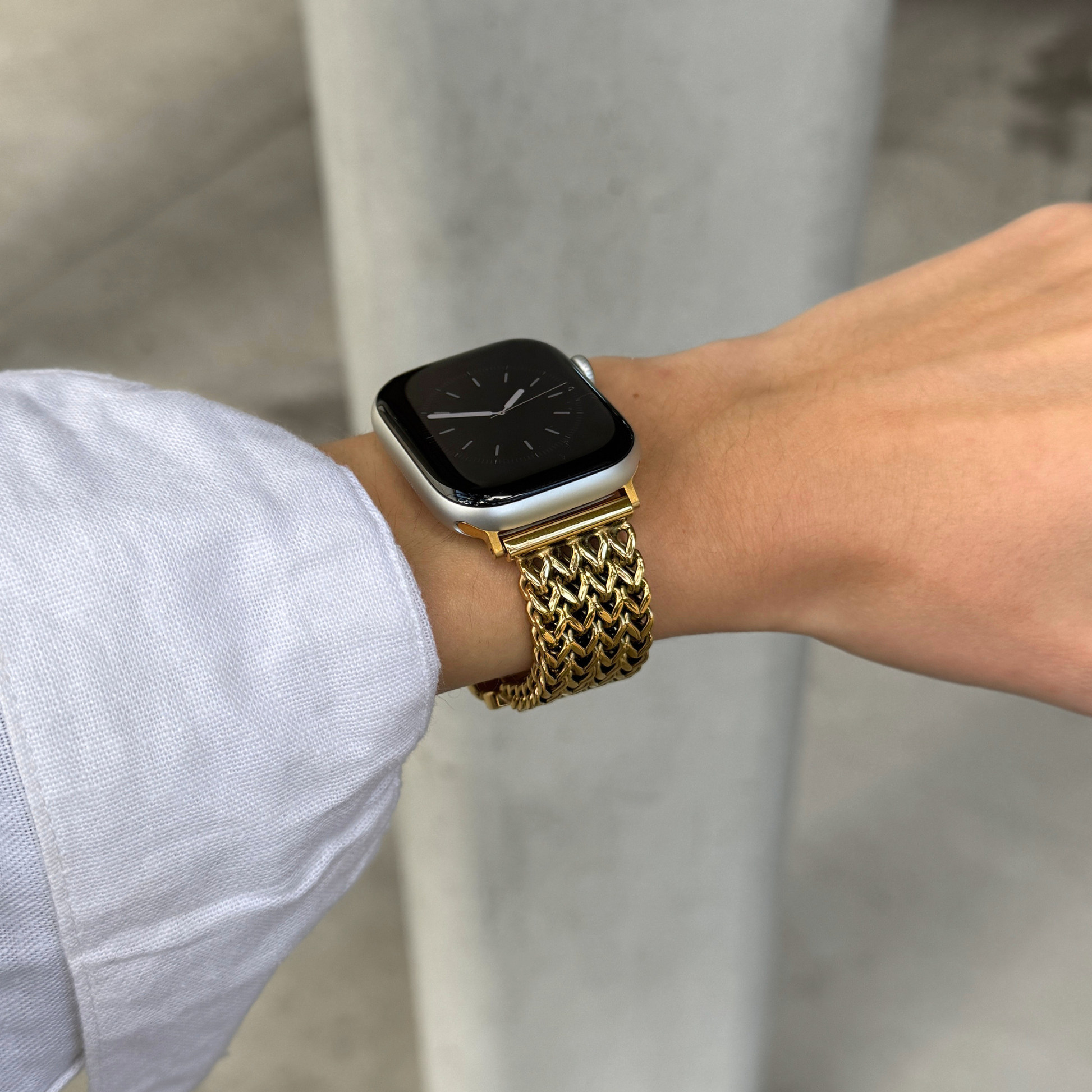 A close-up of a person's wrist wearing a gold-tone metal mesh watch band on an Apple Watch, with a light blue shirt sleeve visible and a blurred gray concrete background.