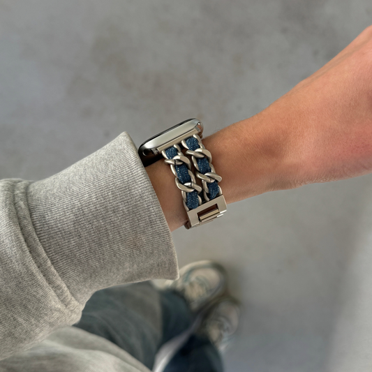 A close-up of a person's wrist wearing a starlight-toned chain-link Apple watch band with blue textured inlay, paired with a gray ribbed sweatshirt and jeans against a concrete floor.